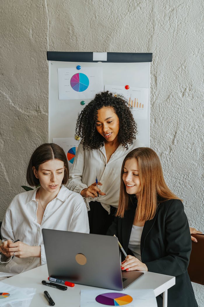 Three businesswomen work together on a laptop in a meeting room with charts.